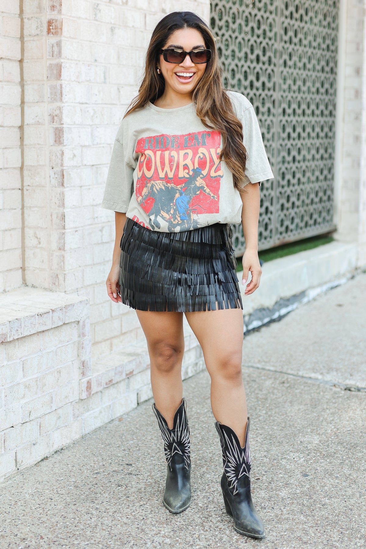 Woman wearing a graphic t-shirt, skirt, and cowboy boots standing against a brick wall.