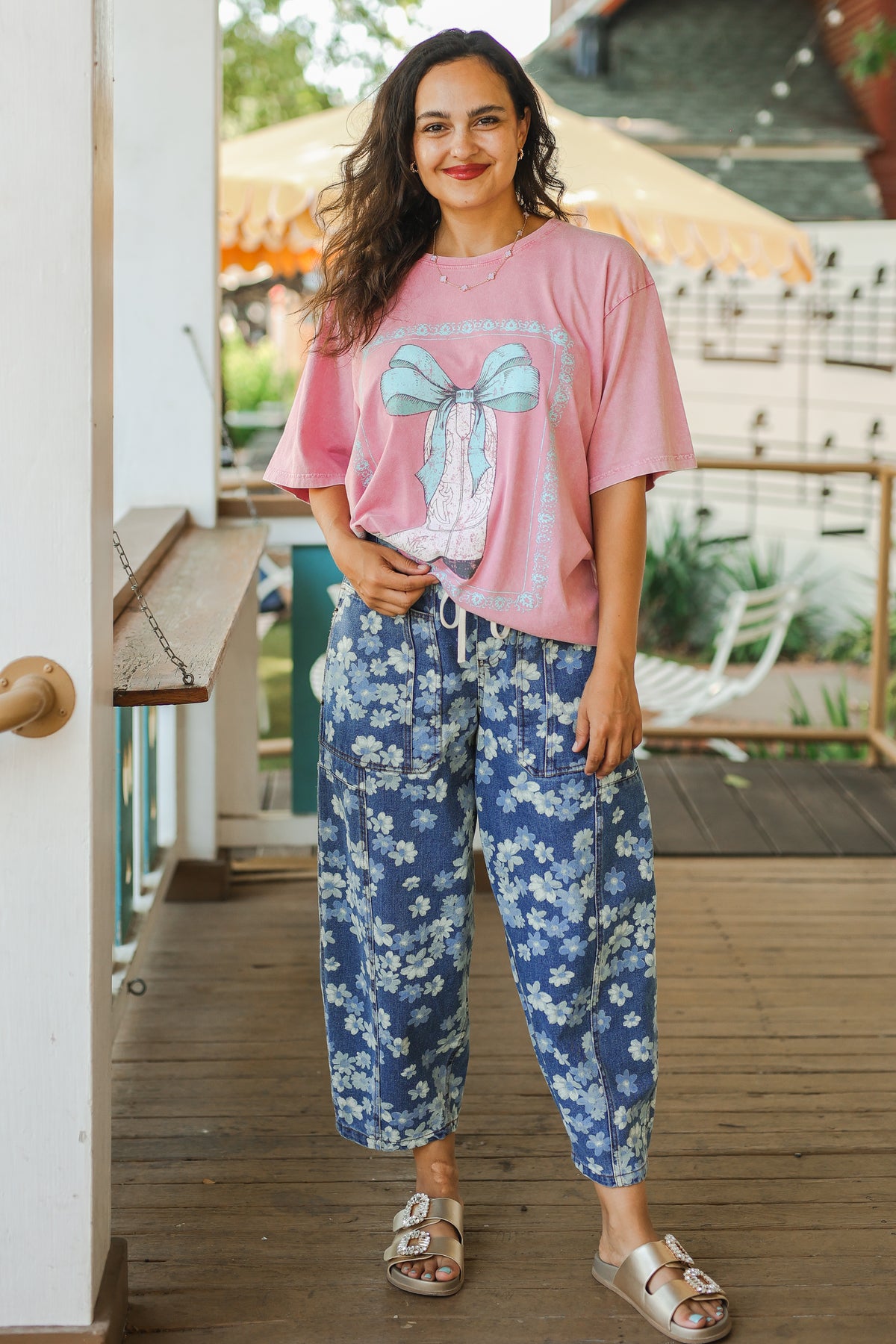 Woman wearing a pink oversized shirt with a bow design and blue floral pants on a wooden deck.