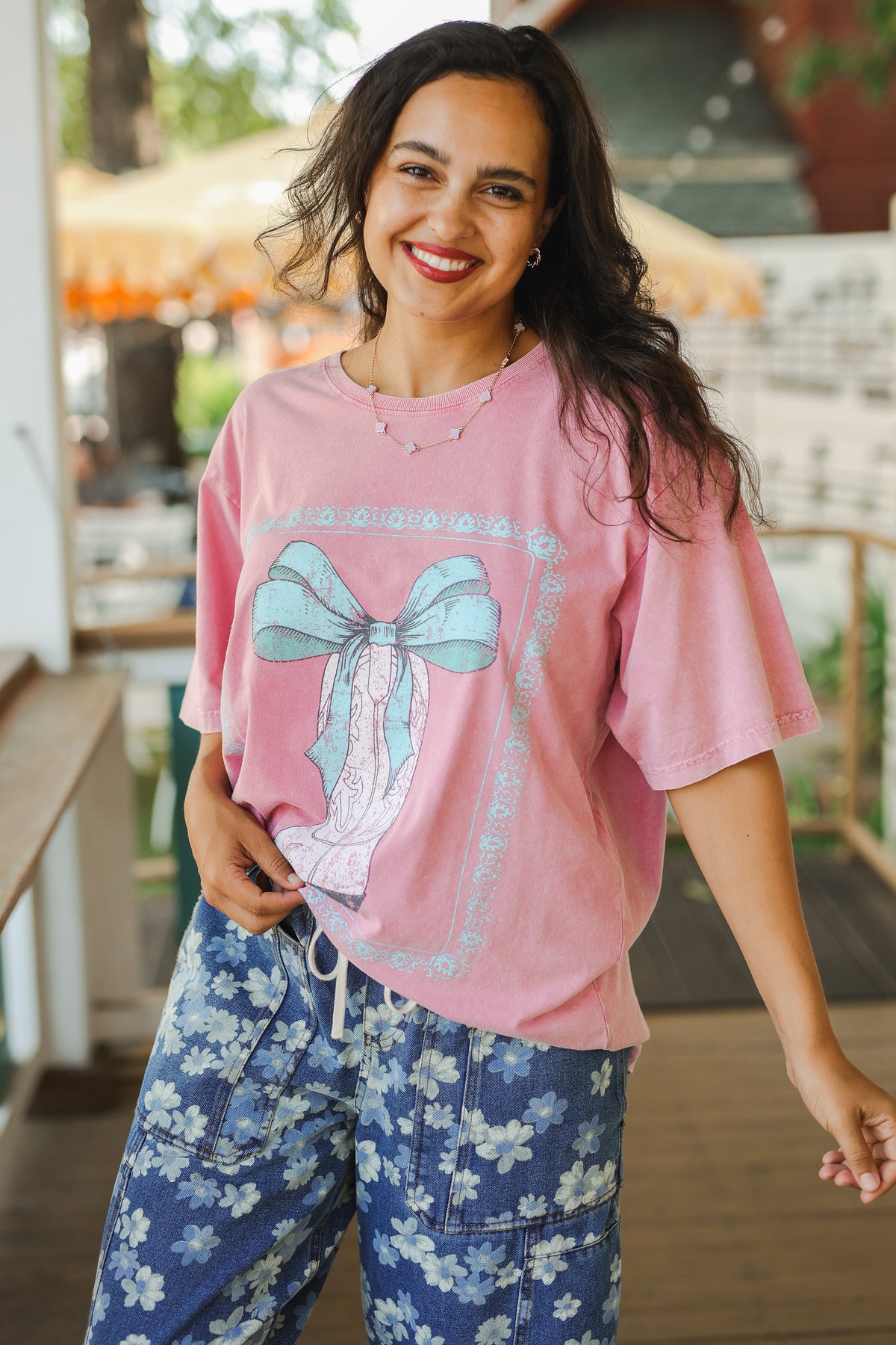 Woman wearing a pink t-shirt with a bow design and blue floral pants outdoors.