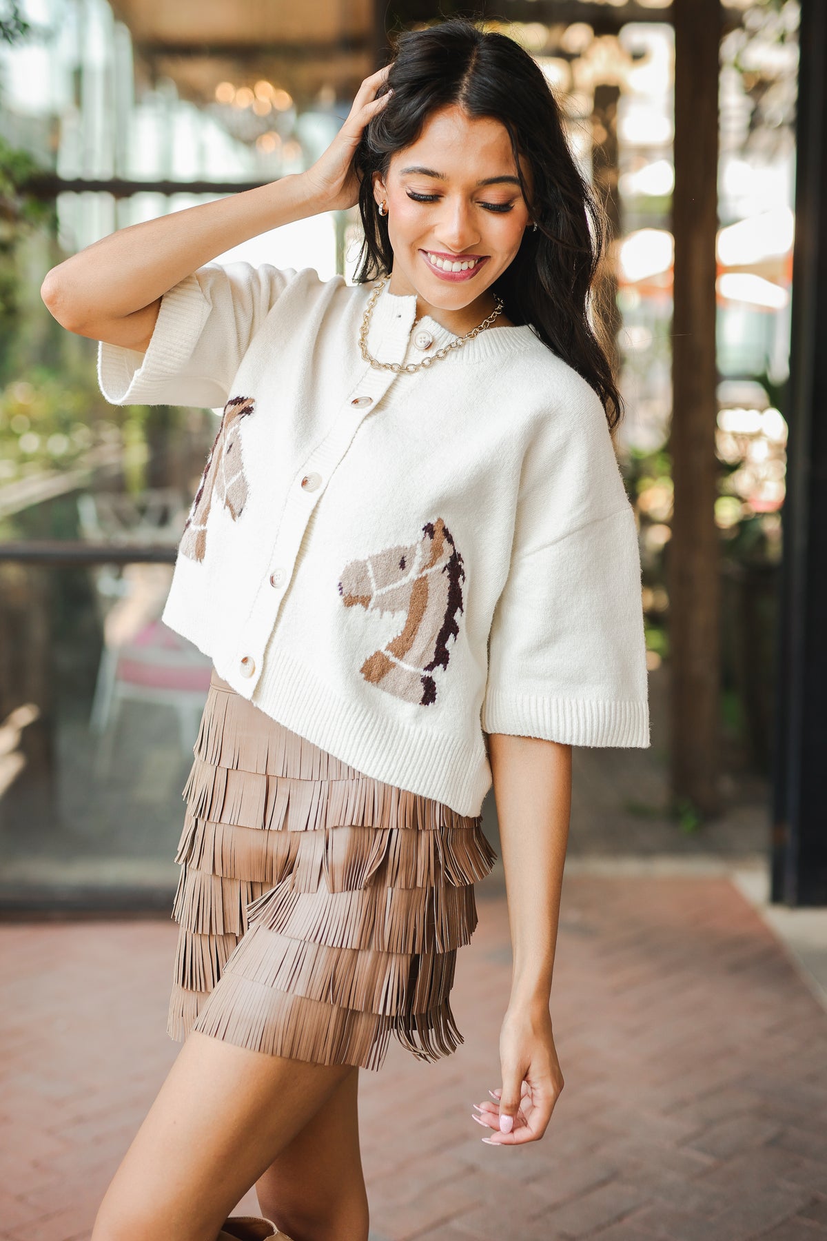 Woman wearing a white sweater with horse designs and a beige fringe skirt outdoors.