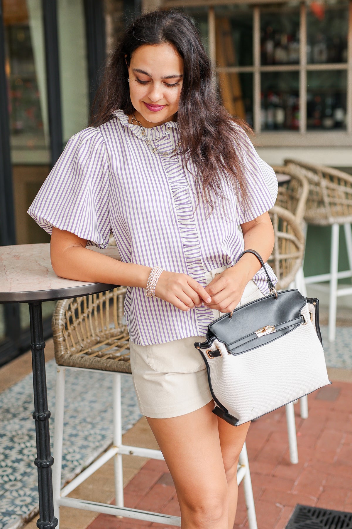 Woman in a striped shirt and shorts holding a black handbag in an outdoor setting.