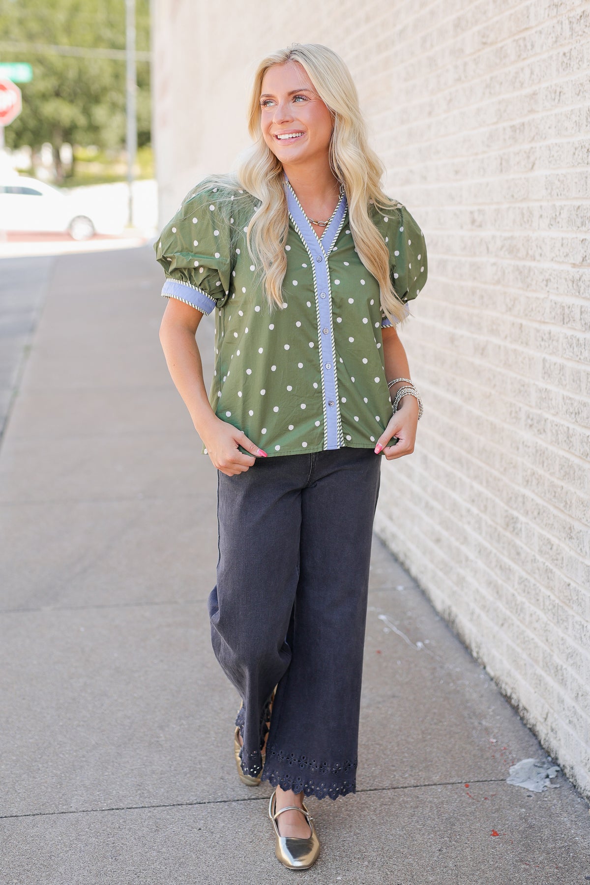 A woman stands on a sidewalk wearing a green puff-sleeve blouse with white polka dots and a light blue striped button placket, paired with dark wide-leg pants and metallic flat shoes, against a white brick wall.