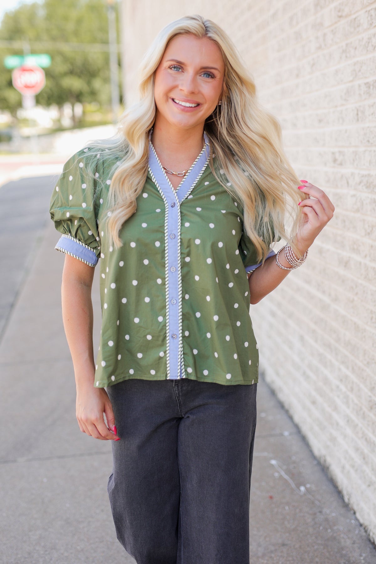 A woman with long blonde hair stands on a sidewalk wearing a green short-sleeve blouse with white polka dots and a light blue patterned trim, paired with dark gray pants.