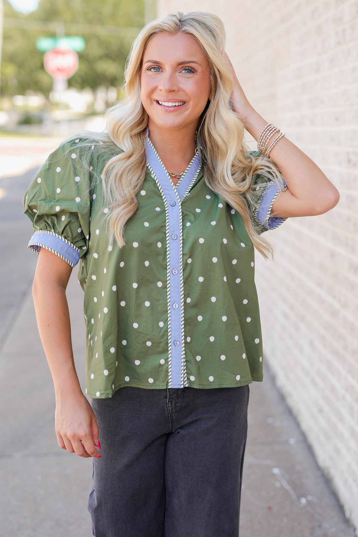 A woman wearing an olive green short-sleeve blouse with white polka dots and light blue trim, paired with dark gray pants, standing outdoors next to a light-colored brick wall.