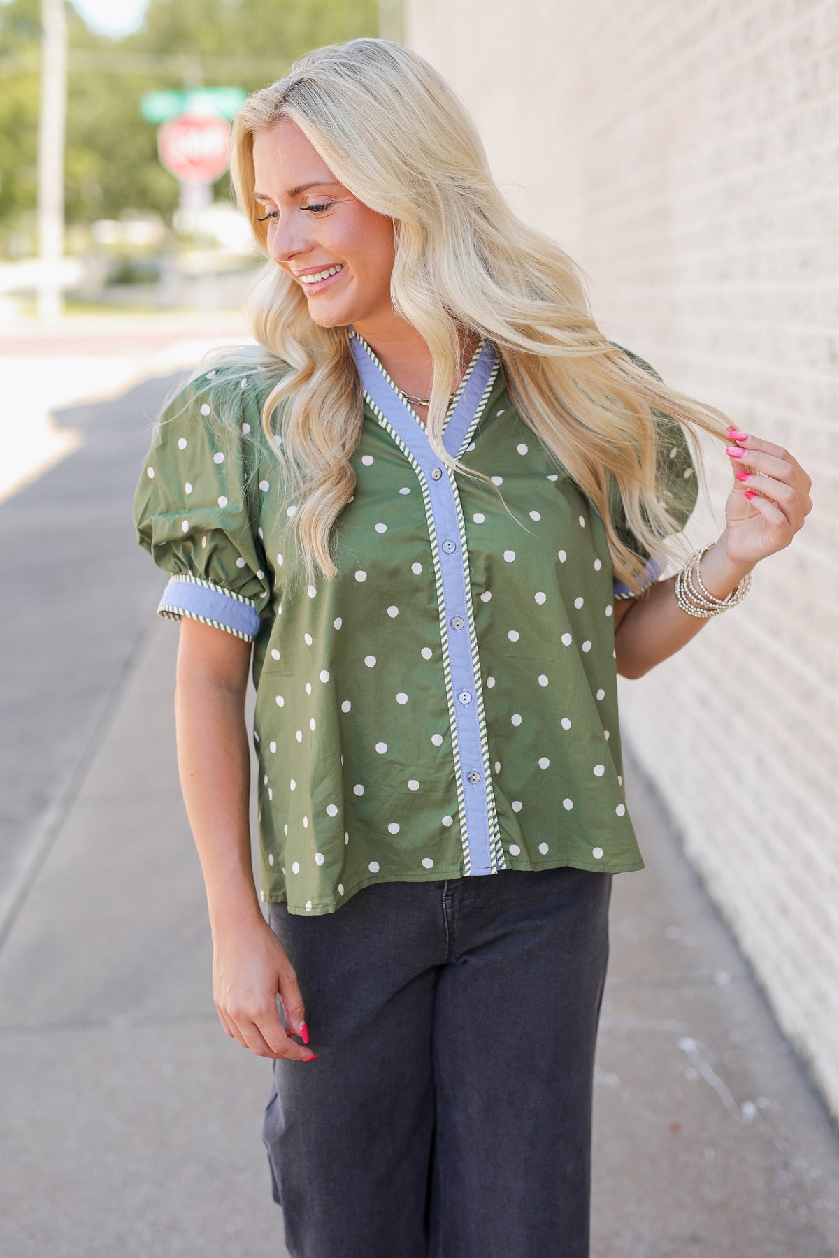 A woman wearing an olive green short-sleeve blouse with white polka dots and light blue trim, paired with dark gray pants, standing outdoors next to a light-colored brick wall.