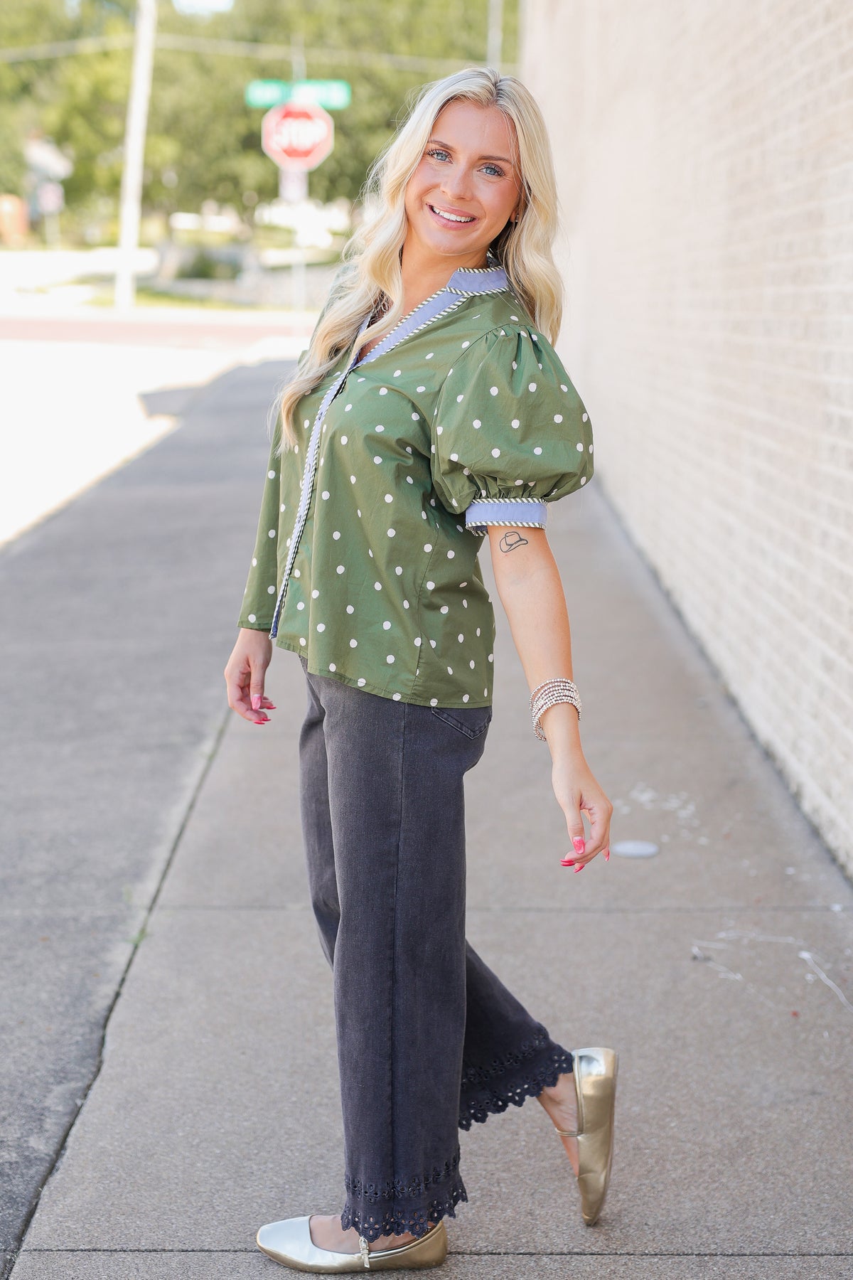 A woman modeling a green short-sleeve blouse with white polka dots and puff sleeves, paired with dark gray wide-leg pants and metallic flats, standing on an outdoor sidewalk.