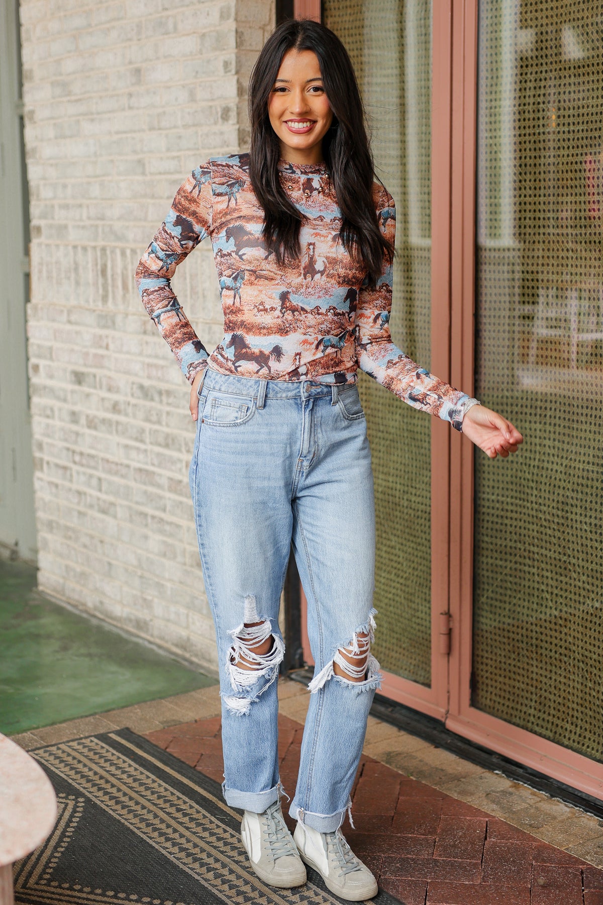 Woman wearing a patterned blouse and ripped jeans standing outside.