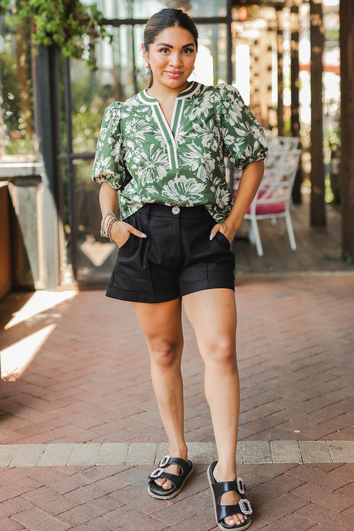 Woman wearing a green floral blouse and black shorts standing on a brick patio.