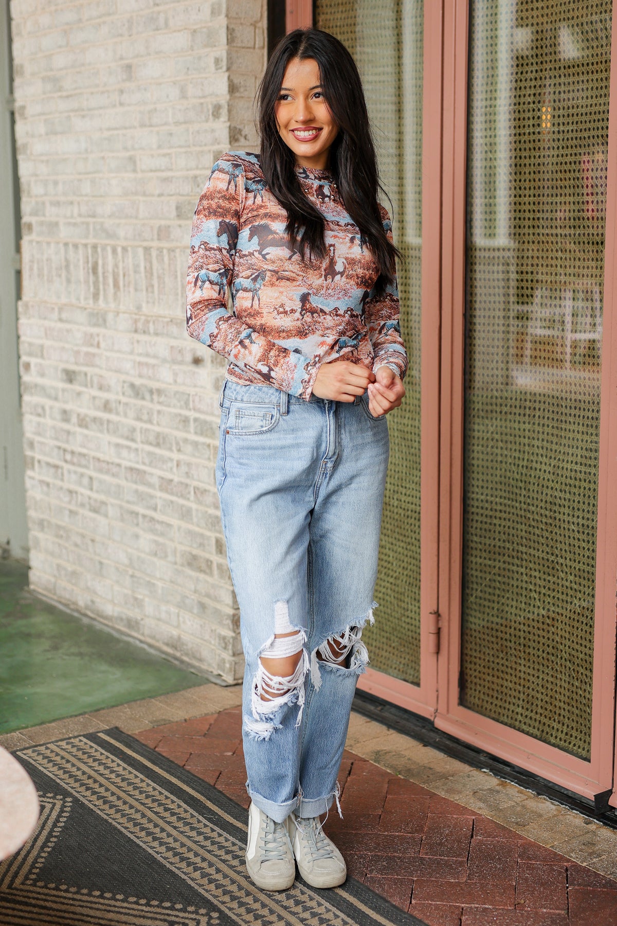 Woman wearing a patterned blouse and ripped jeans standing in front of a building entrance.
