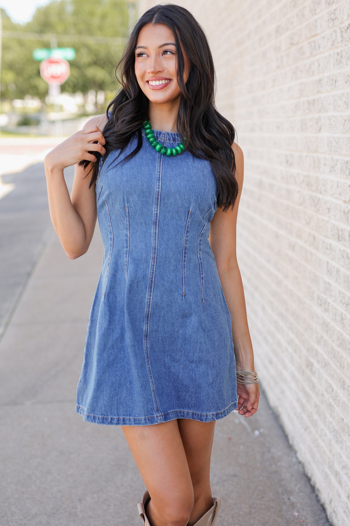 A woman wearing a sleeveless blue denim dress and a chunky green beaded necklace, standing outdoors against a white brick wall.