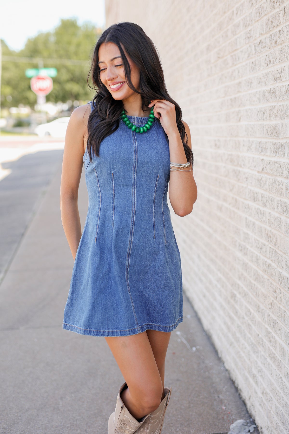 A woman wearing a sleeveless blue denim dress and beige cowboy boots, standing outdoors next to a light-colored brick wall.