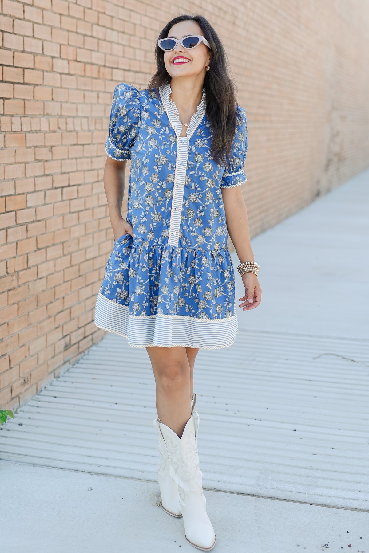 Woman wearing a blue floral dress with white boots against a brick wall.