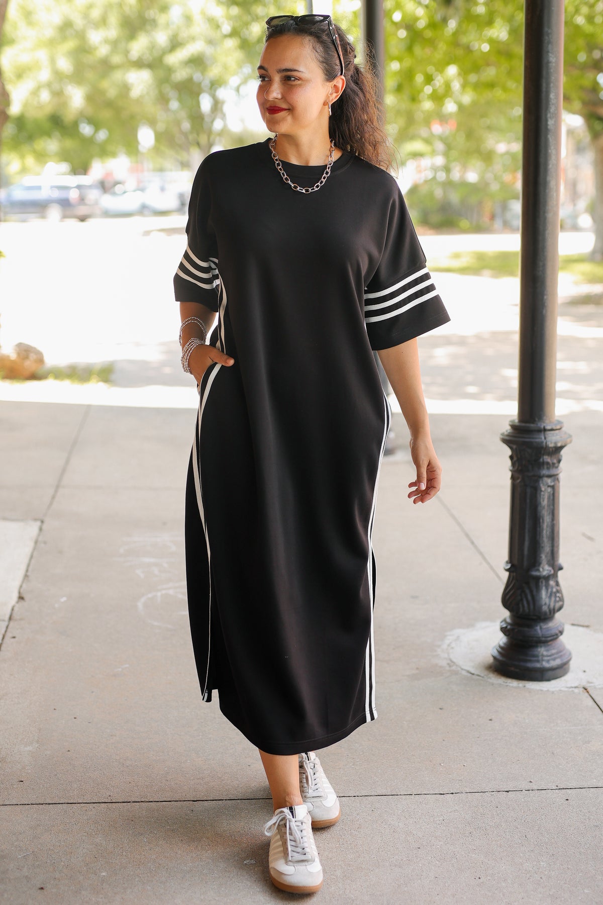 A woman wearing a black midi dress with white stripes on the sleeves and sides, styled with white sneakers, standing on an outdoor sidewalk next to a black metal pole.