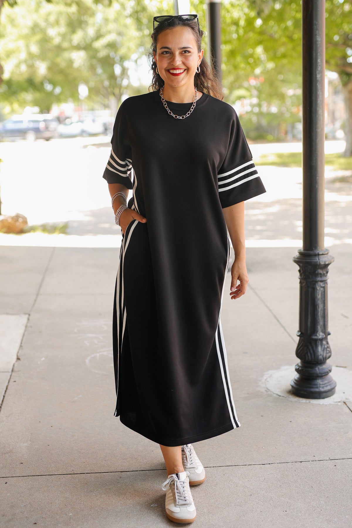 A woman modeling a black midi dress with white stripe details on the sleeves and sides, paired with white sneakers, standing on a sidewalk under a black metal column.