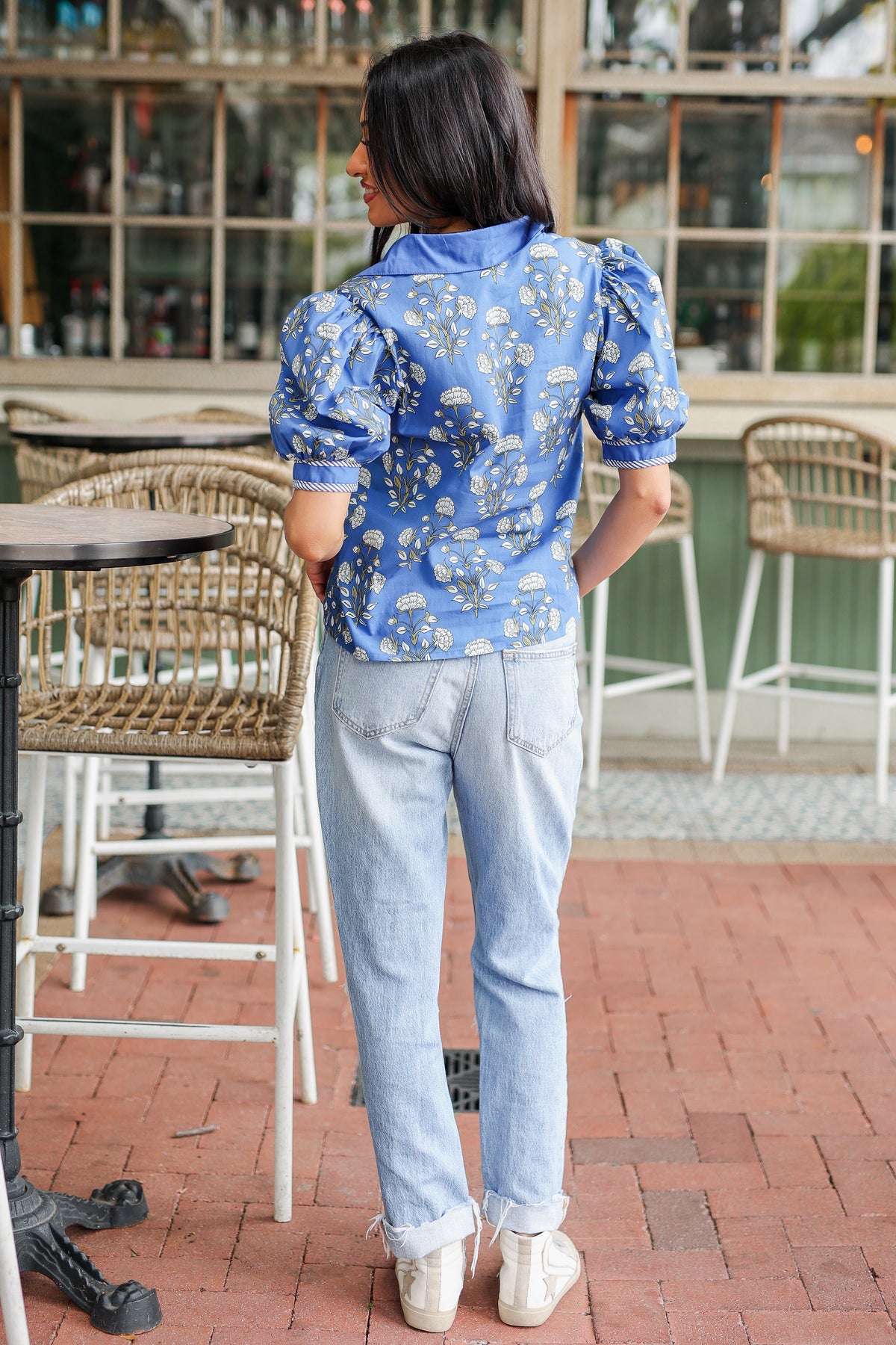 Woman in a blue floral blouse and light jeans standing on a patio.