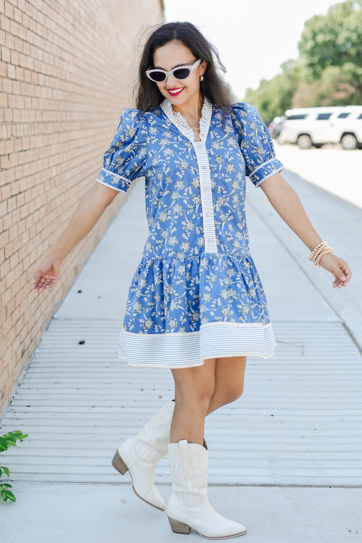 Woman in a blue floral dress with white boots standing against a brick wall.