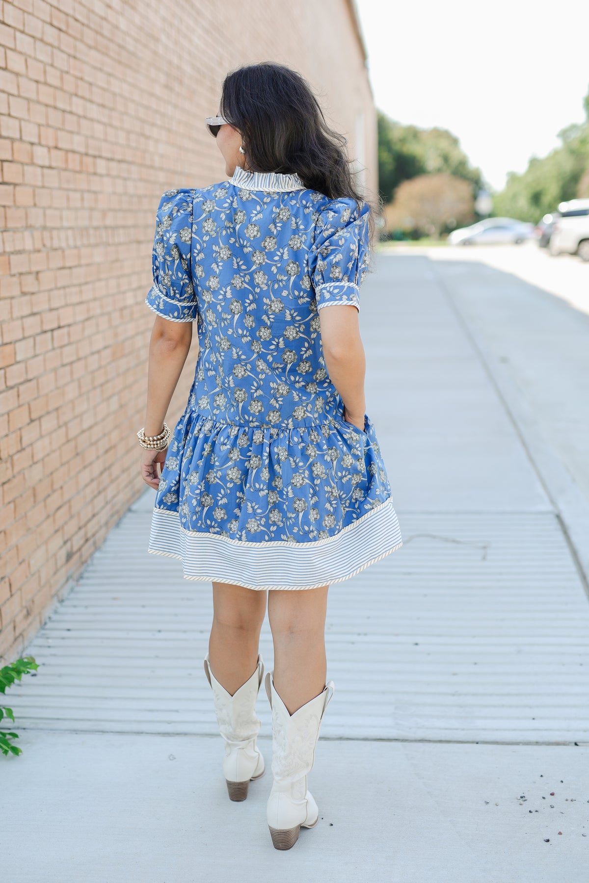 Woman in a blue floral dress and white cowboy boots walking on a sidewalk.