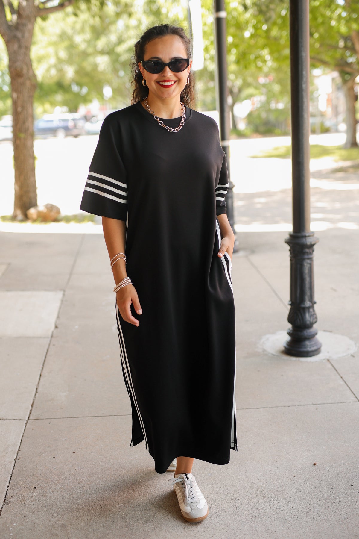 A woman wearing a black midi dress with white stripes on the sleeves and sides, styled with white sneakers and sunglasses, standing on an outdoor sidewalk.