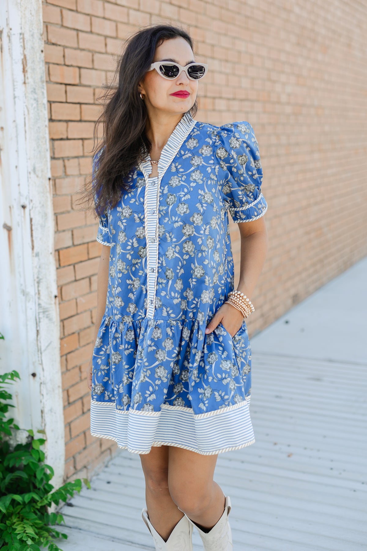 Woman wearing a blue floral dress with white boots against a brick wall.