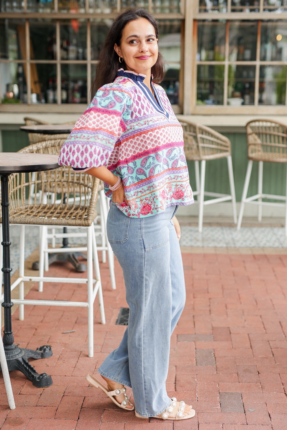 Woman in a colorful patterned blouse and jeans standing outdoors on a patio.