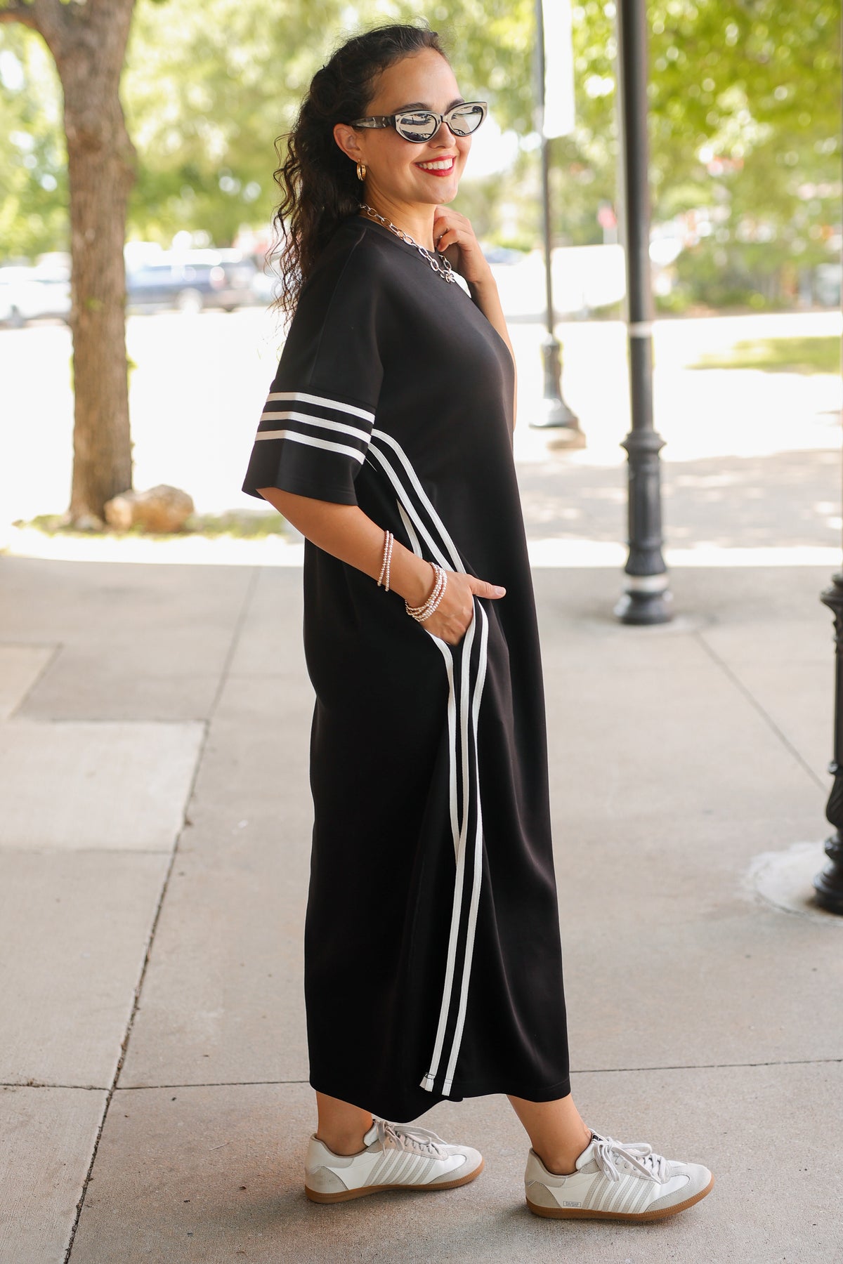 A woman wearing a black midi dress with white stripes on the sleeves and side, styled with white sneakers and sunglasses, standing on a sidewalk.