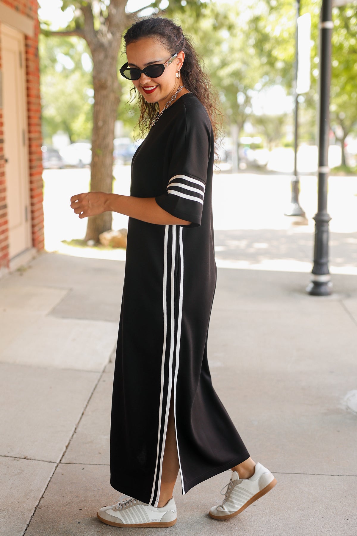 A woman wearing a black maxi dress with white stripes on the sleeves and down the side, styled with white sneakers and sunglasses, walking on a sunny outdoor sidewalk.