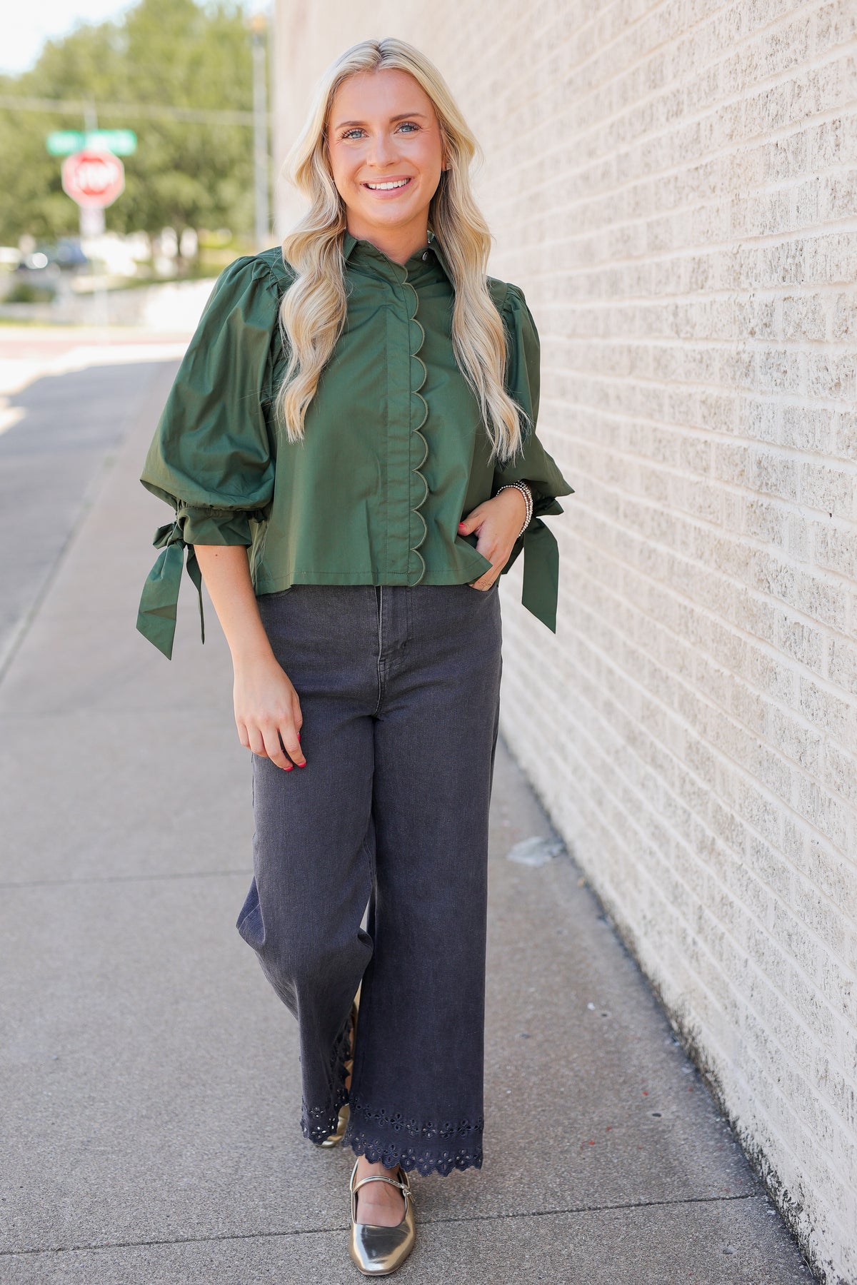 A woman stands on a sidewalk next to a light brick wall, wearing an olive green ruffled blouse with puffed sleeves and dark charcoal wide-leg pants.