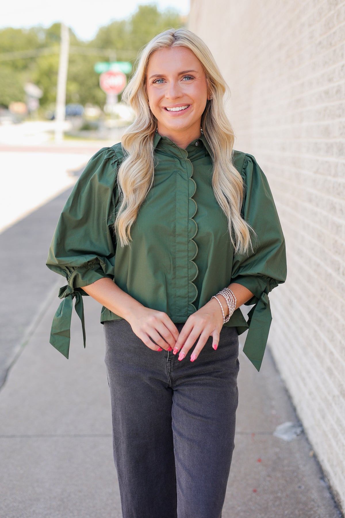 A smiling blonde woman wearing an olive green long-sleeve blouse with puffed sleeves, a scalloped front placket, and side tie details, paired with dark gray jeans, standing outdoors next to a light brick wall.