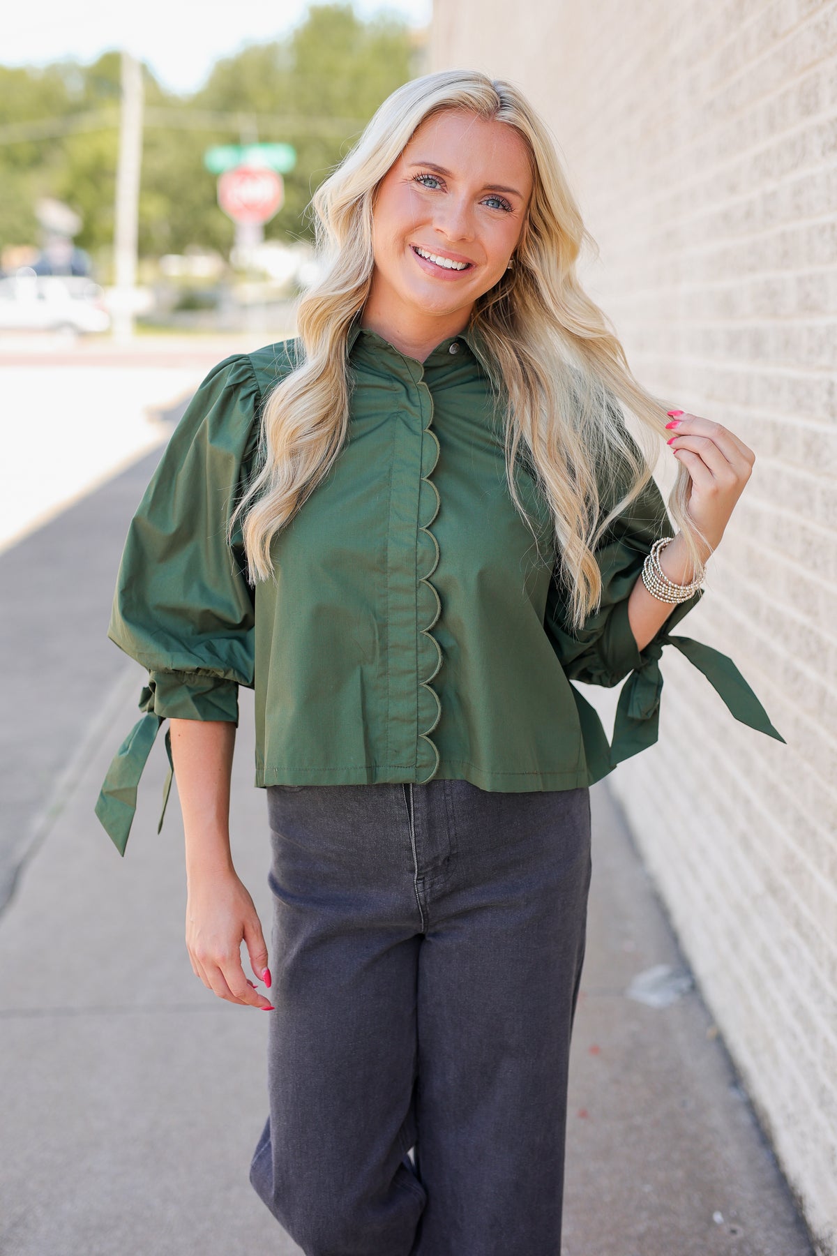 A woman with long blonde hair stands outdoors against a light brick wall wearing a dark green puff-sleeve blouse with scalloped front trim and dark charcoal gray pants.