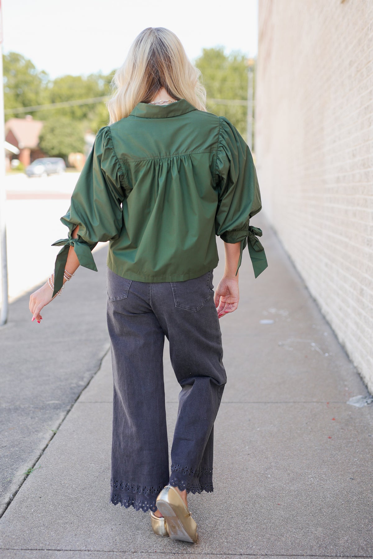 A woman viewed from behind walking on a sidewalk, wearing an olive green satin blouse with puffed sleeves and tie details, dark gray wide-leg jeans, and gold shoes.