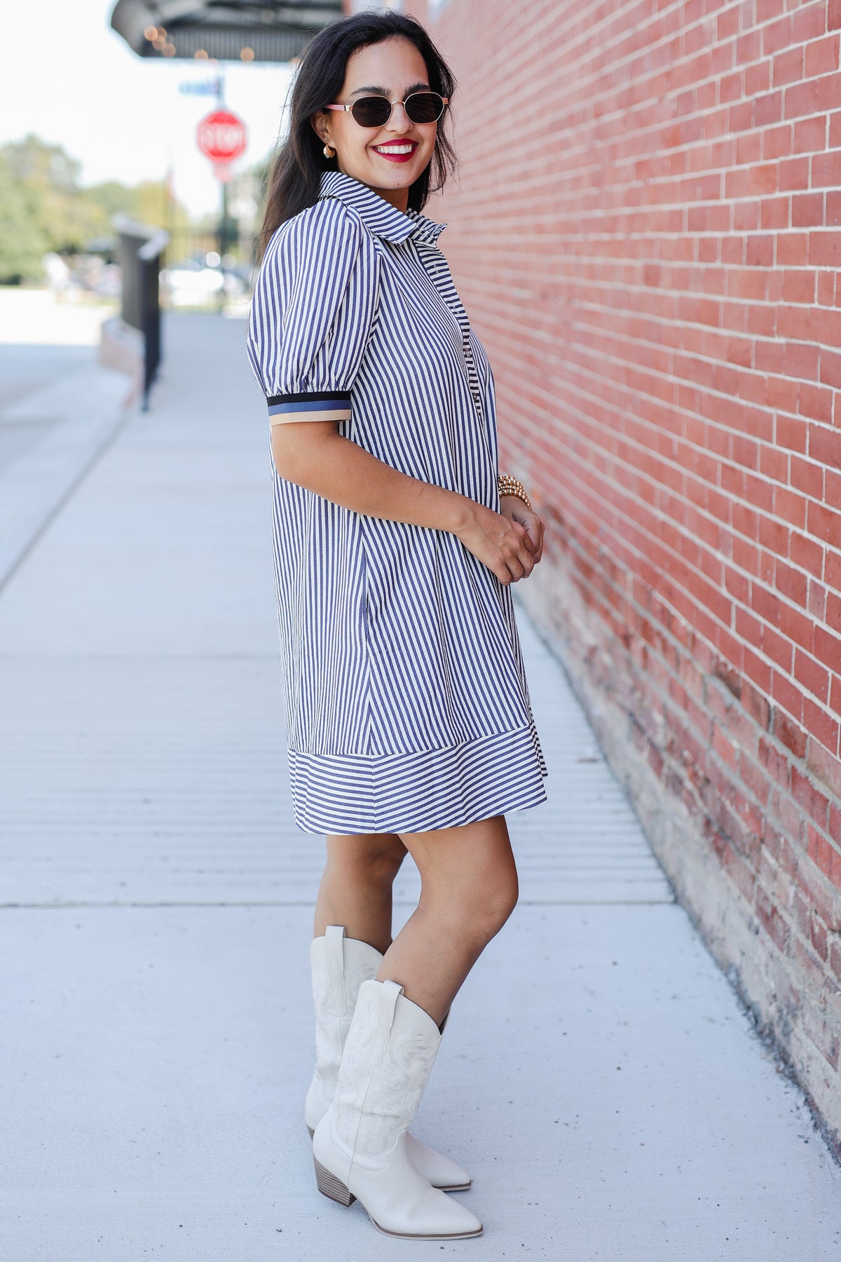 Woman wearing a blue and white striped dress with white boots against a brick wall.