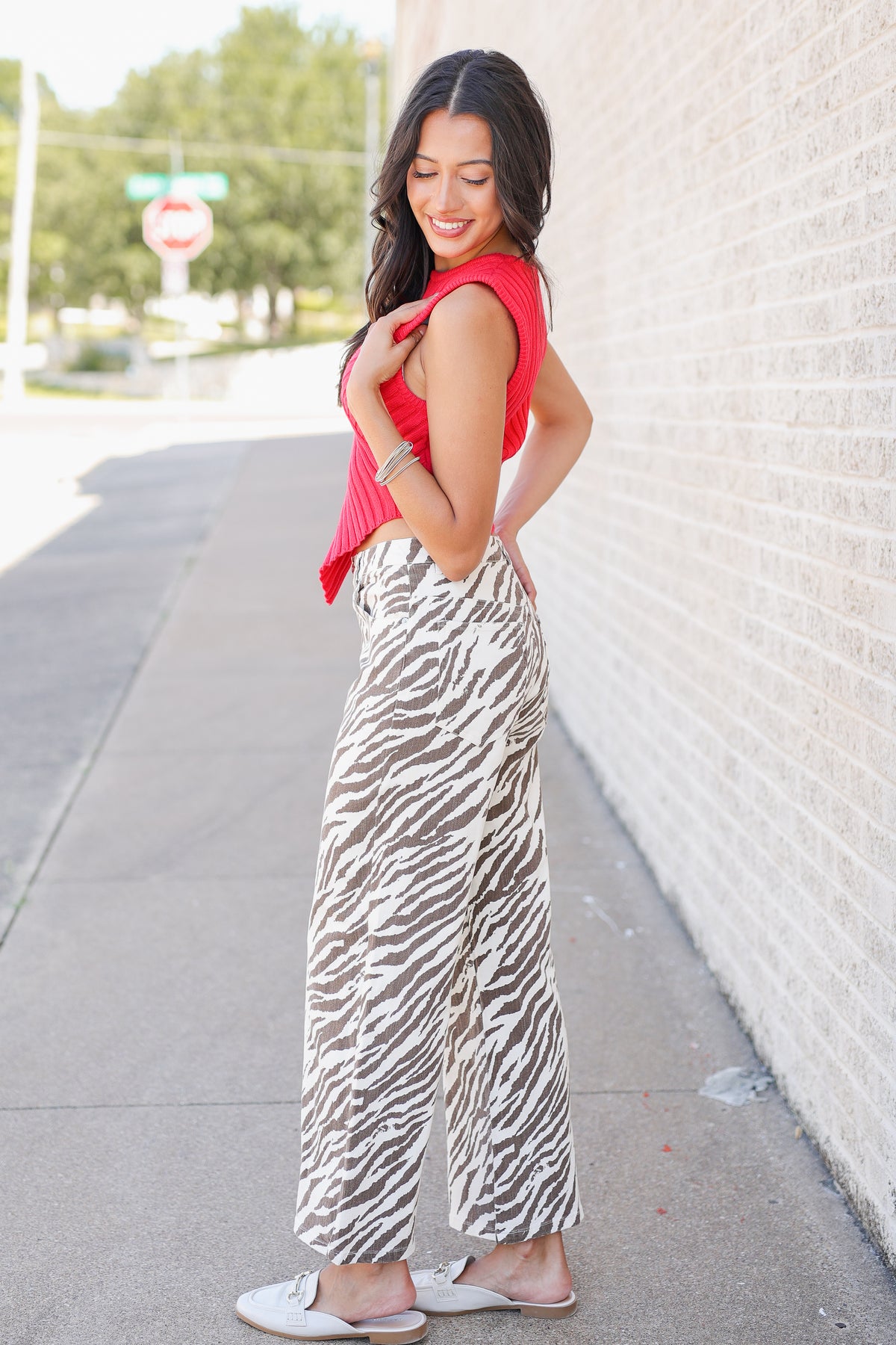 A woman posing outdoors on a sidewalk next to a white brick wall, wearing a red sleeveless top and high-waisted zebra-print wide-leg pants.