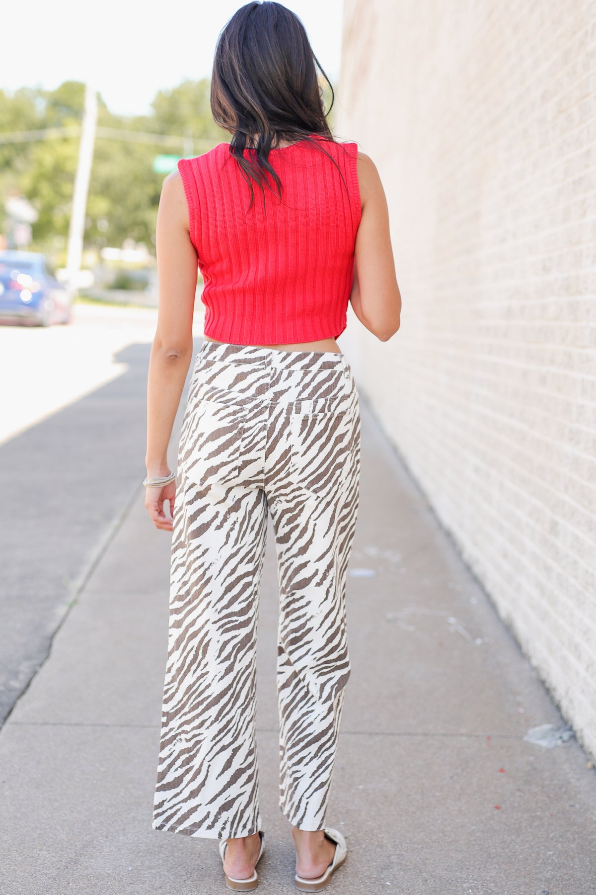 A woman viewed from behind wearing a bright red ribbed sleeveless top and beige-and-white zebra-print wide-leg pants, standing on a sidewalk next to a light brick wall.