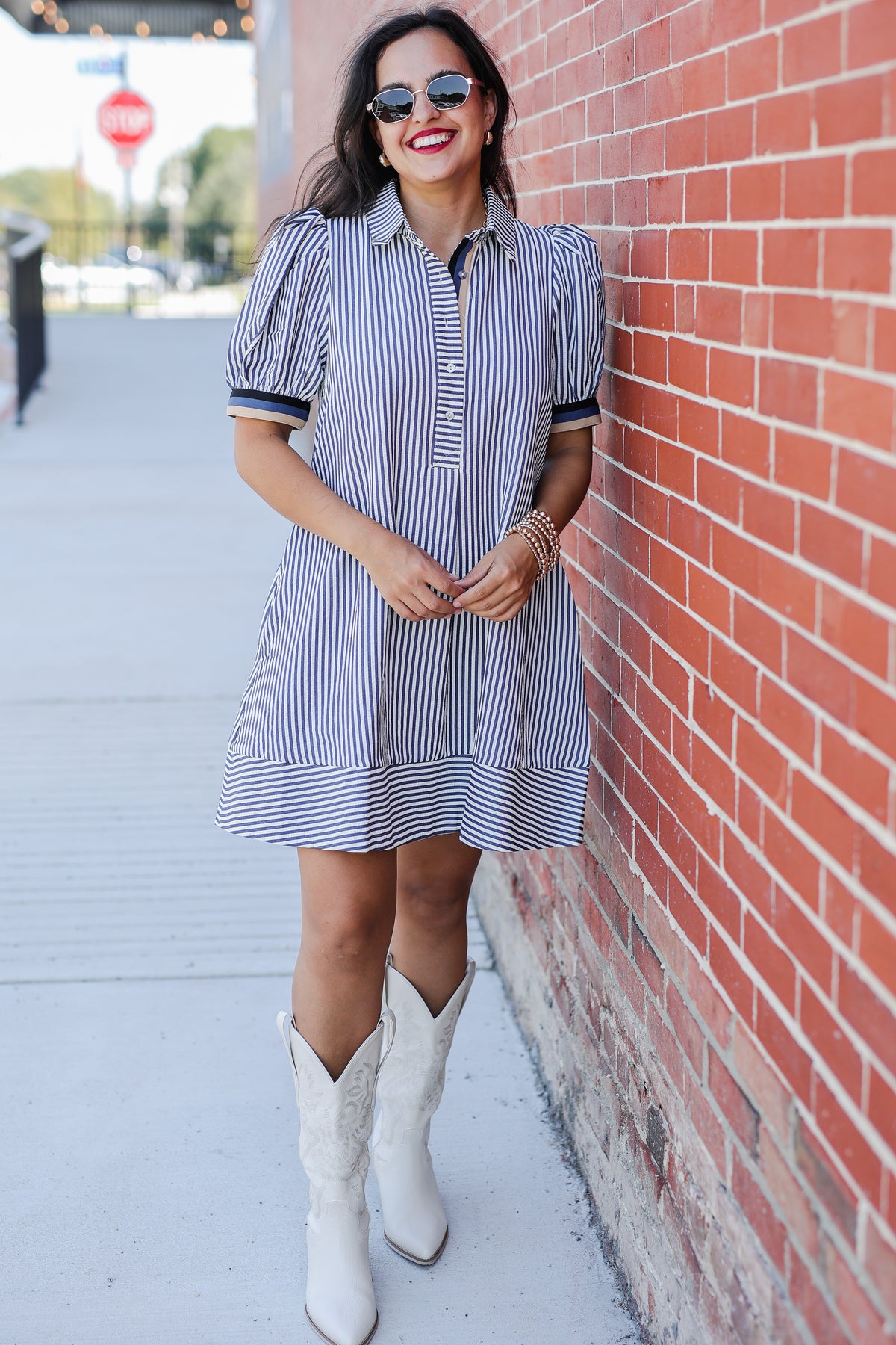Woman in a striped dress and white boots leaning against a brick wall.