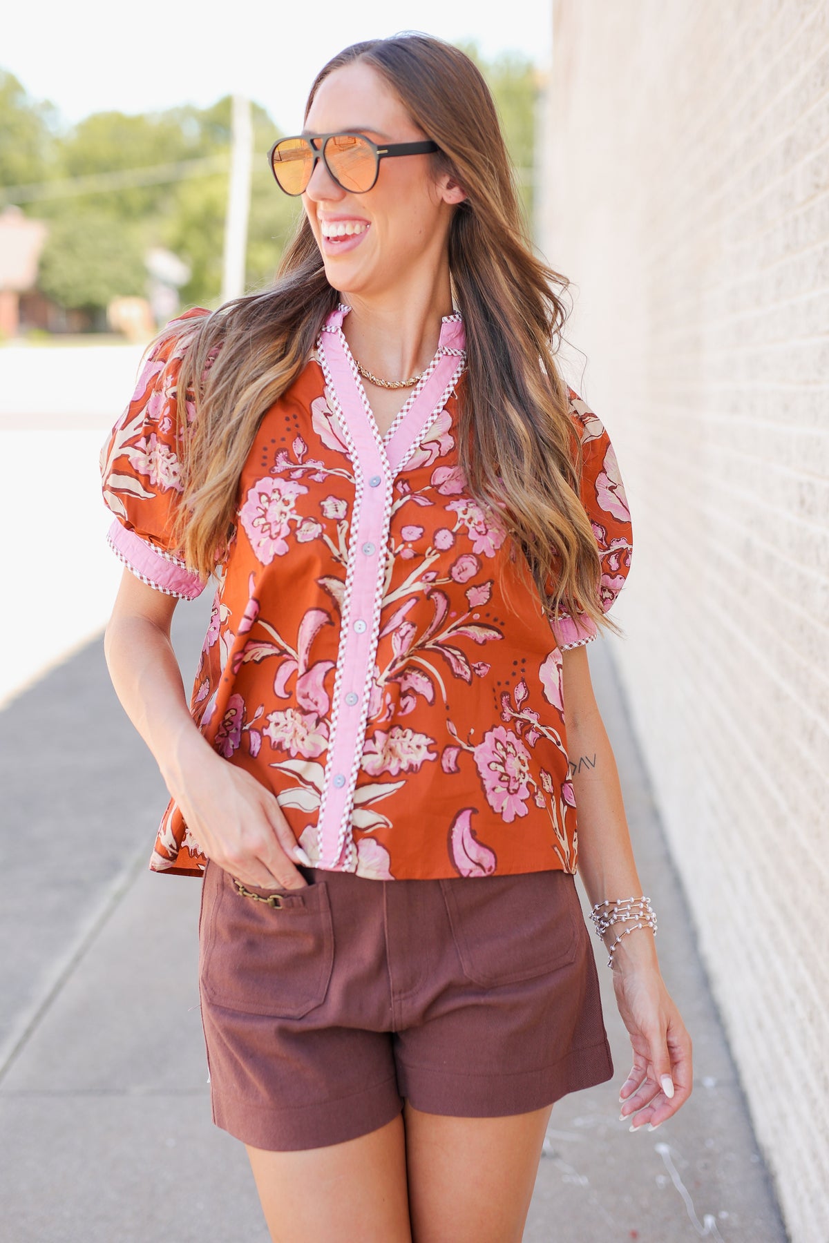 A woman wearing a rust-orange short-sleeve button-up shirt with a pink floral print and pink trim, paired with brown shorts, standing outdoors next to a light-colored brick wall.