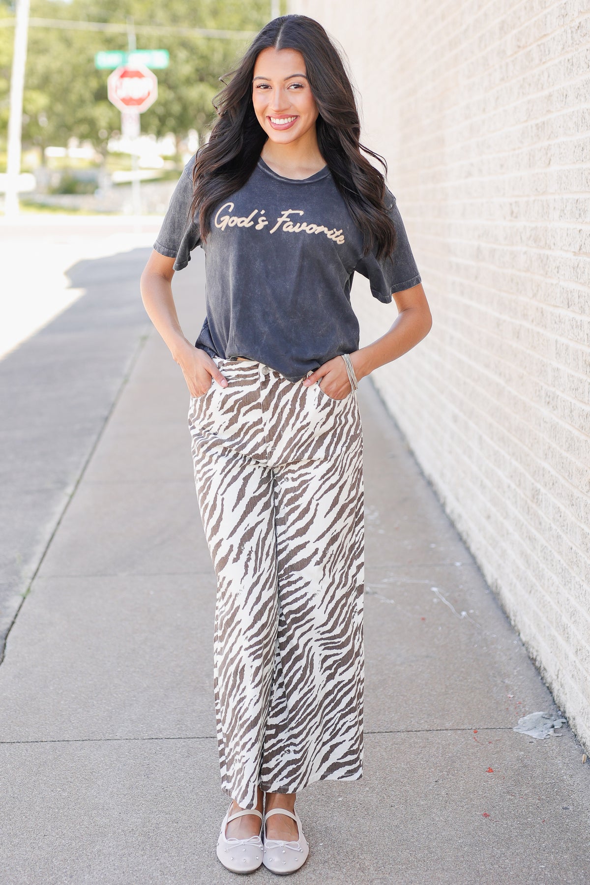 A woman standing on a sidewalk wearing a dark gray graphic t-shirt with 'God's Favorites' printed on the chest and high-waisted, wide-leg zebra-print pants in brown and white.