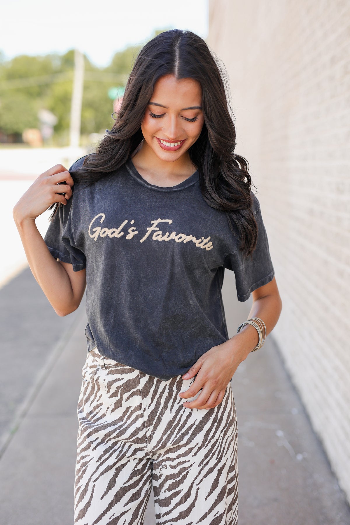 A woman wearing a dark gray washed t-shirt with 'God's Favorites' printed in white script, paired with brown and white zebra-print pants, standing outdoors against a light brick wall.