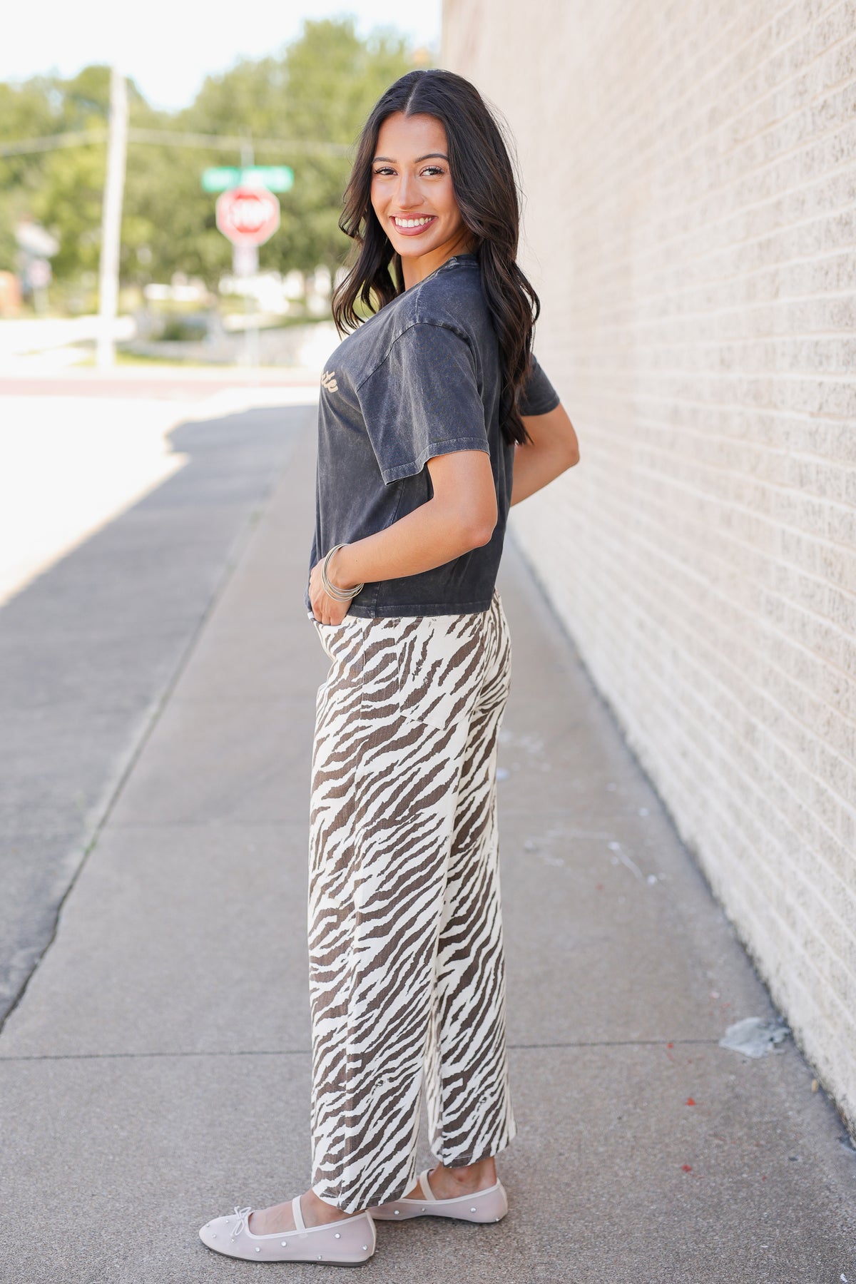 A woman stands on a sidewalk next to a light brick wall, wearing a faded black t-shirt and a long, wide-leg zebra-print skirt with side slits, paired with beige flats.