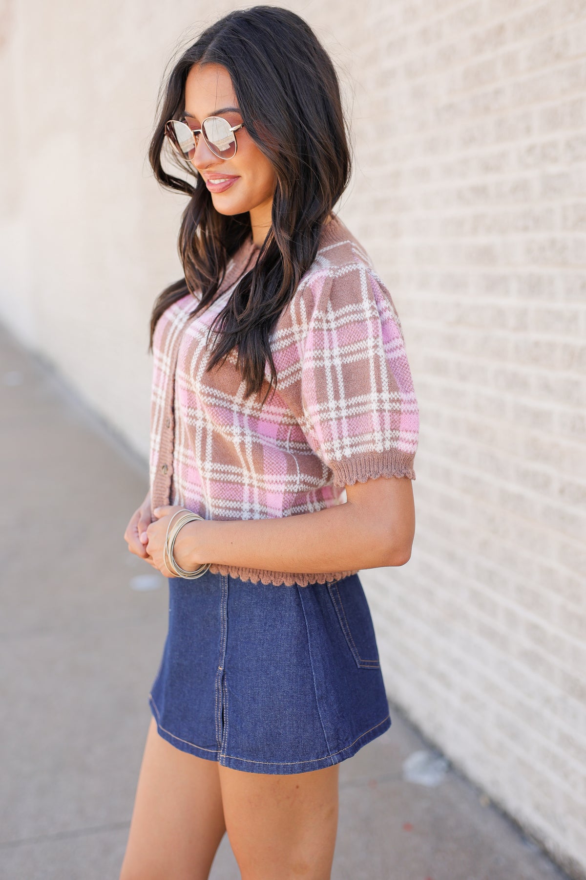 A woman wearing a pink and beige plaid short-sleeve top with a scalloped hem and a blue denim mini skirt, standing outdoors against a light brick wall.