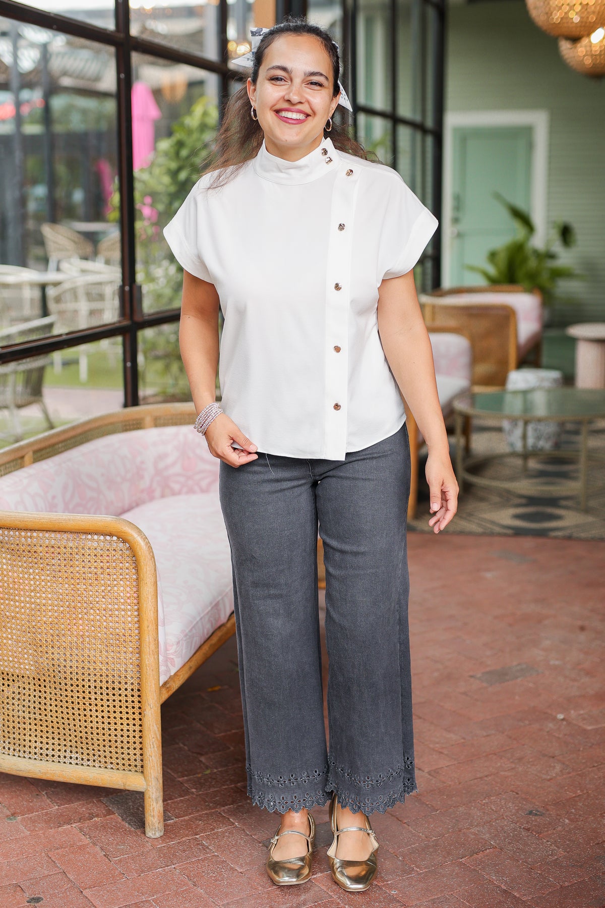 Woman wearing a white blouse and gray pants standing in an outdoor setting with furniture and plants.
