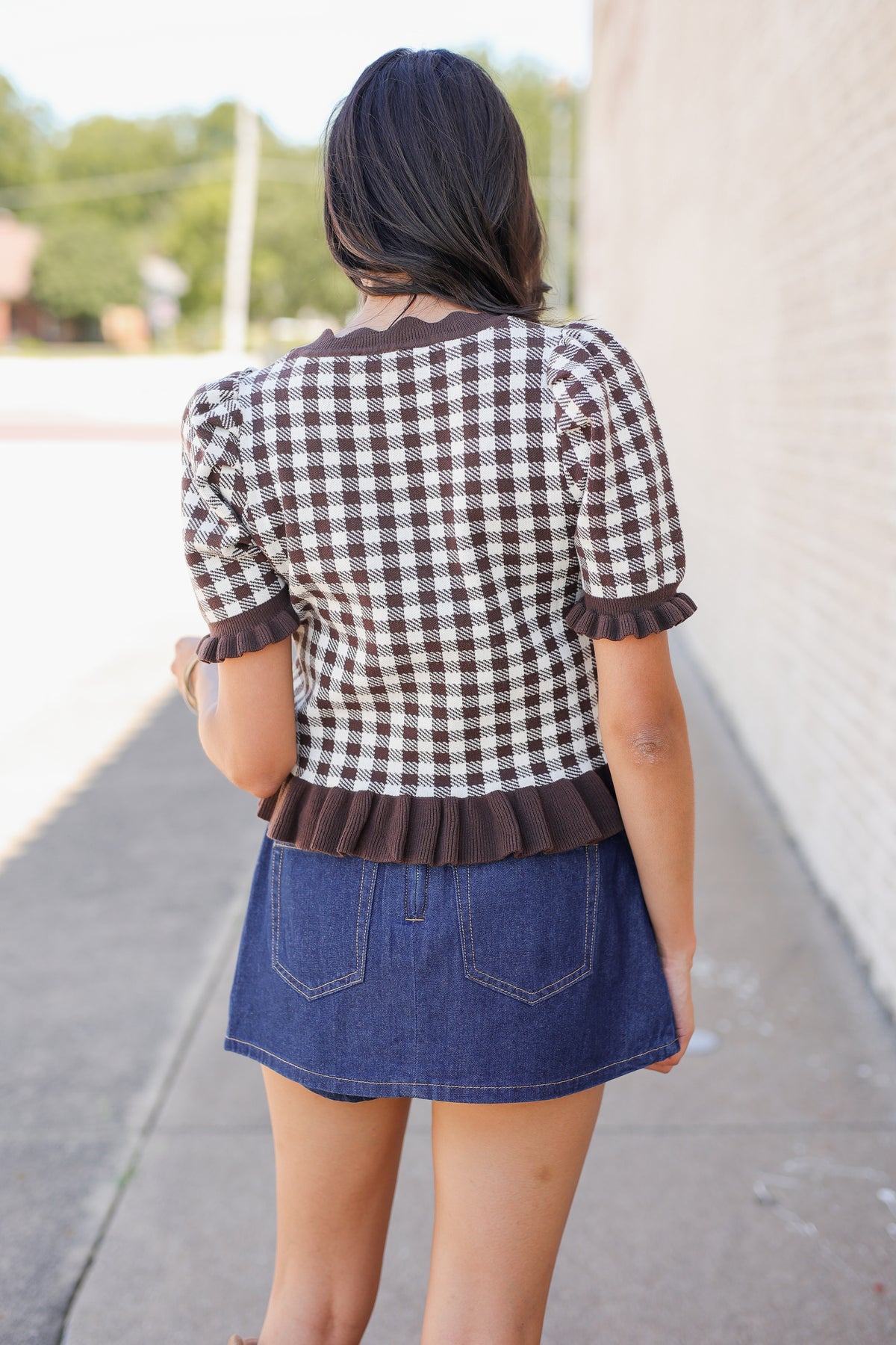 A woman viewed from behind wearing a brown and white gingham short-sleeve top with ruffle trim and a blue denim mini skirt, standing on a sidewalk next to a light-colored brick wall.