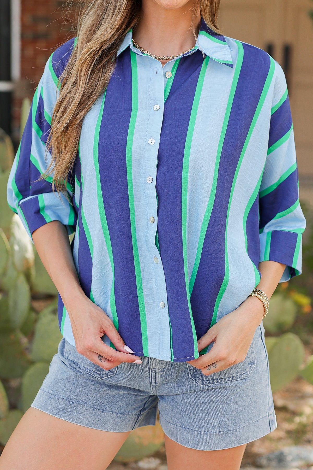 A woman wearing a loose button-front striped blouse with vertical blue, light blue, and green stripes, paired with denim shorts, photographed outdoors with blurred cactus and building elements in the background.
