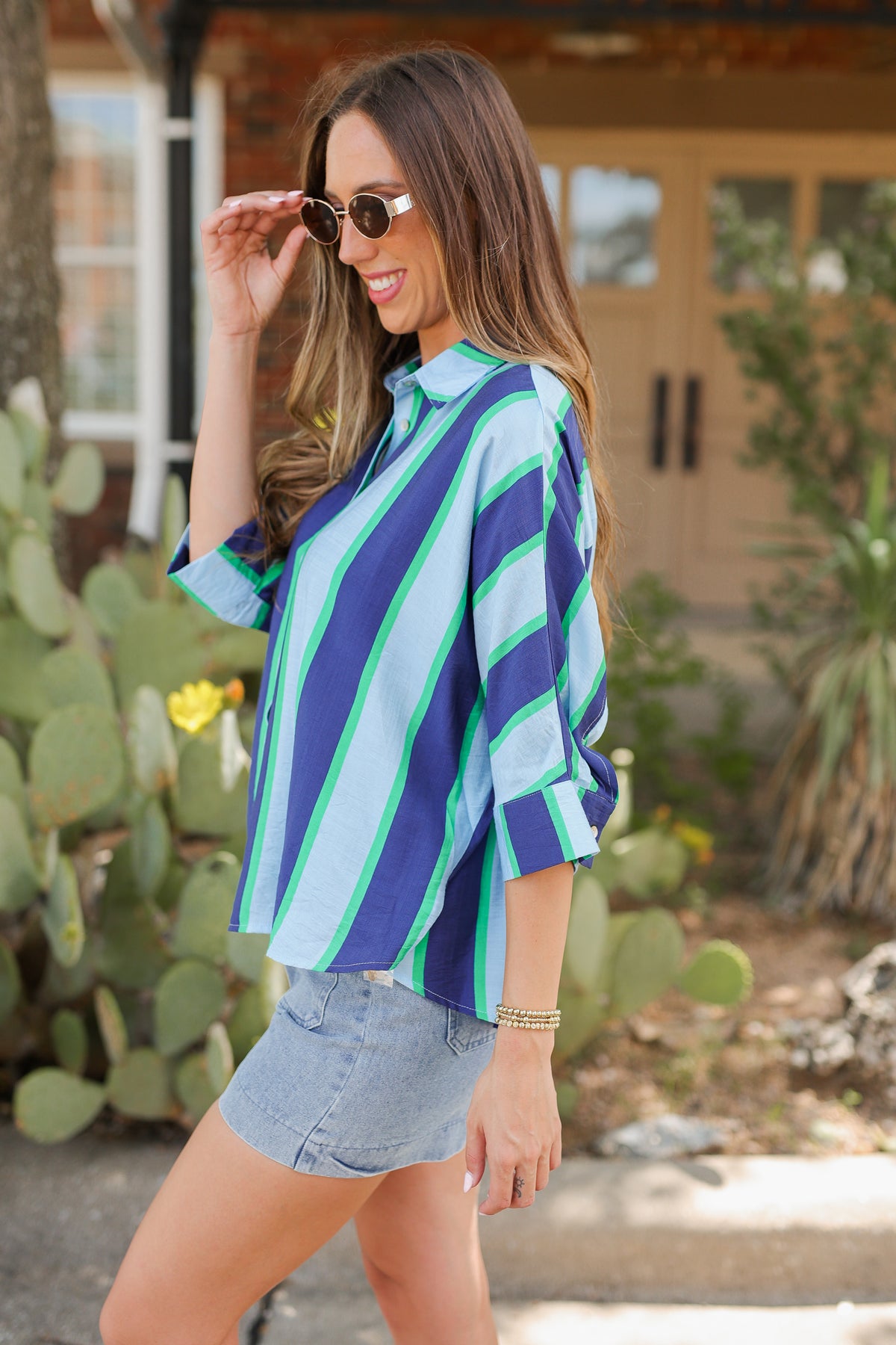 A woman wearing a blue and green striped button-up shirt and denim shorts, standing outdoors in front of a house with cacti in the background.