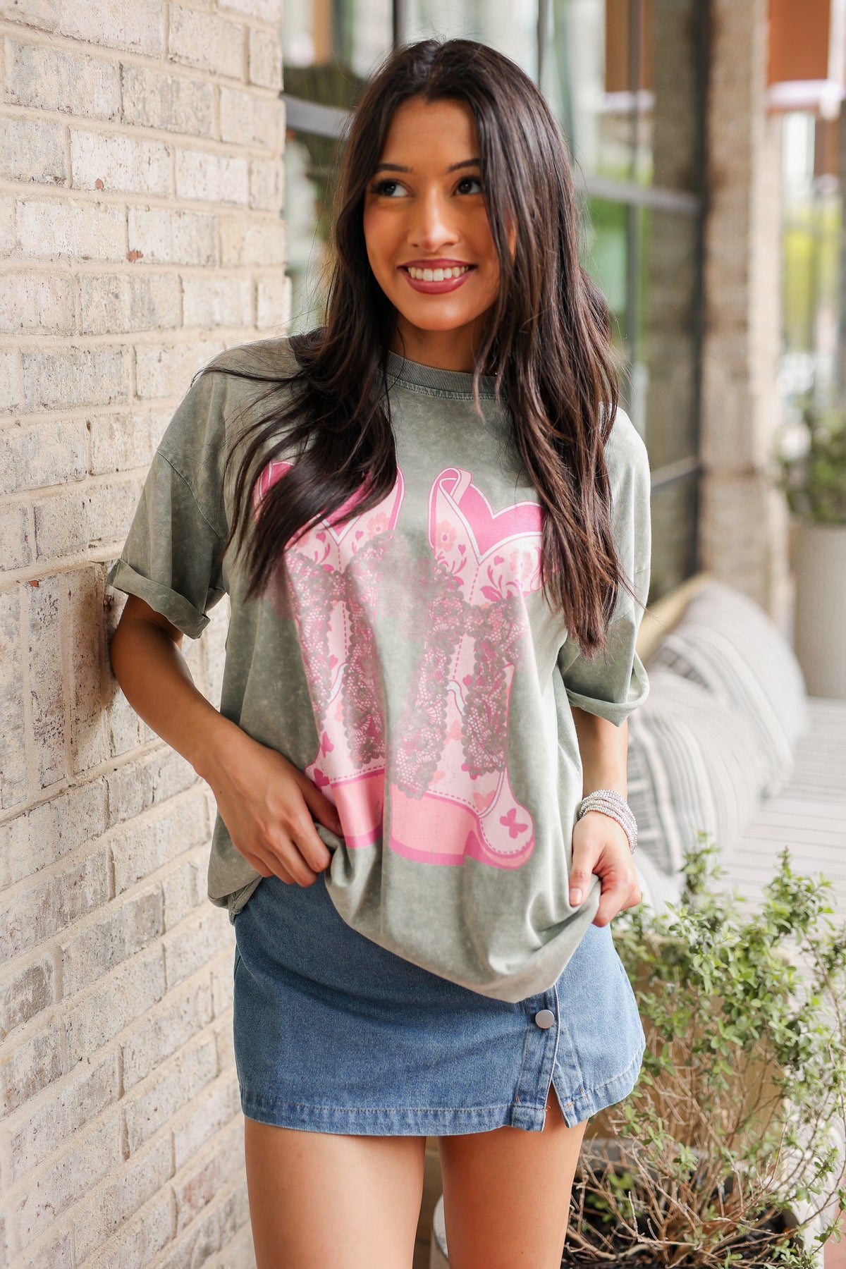 Woman wearing a gray t-shirt with pink graphic design and denim shorts, standing against a brick wall.