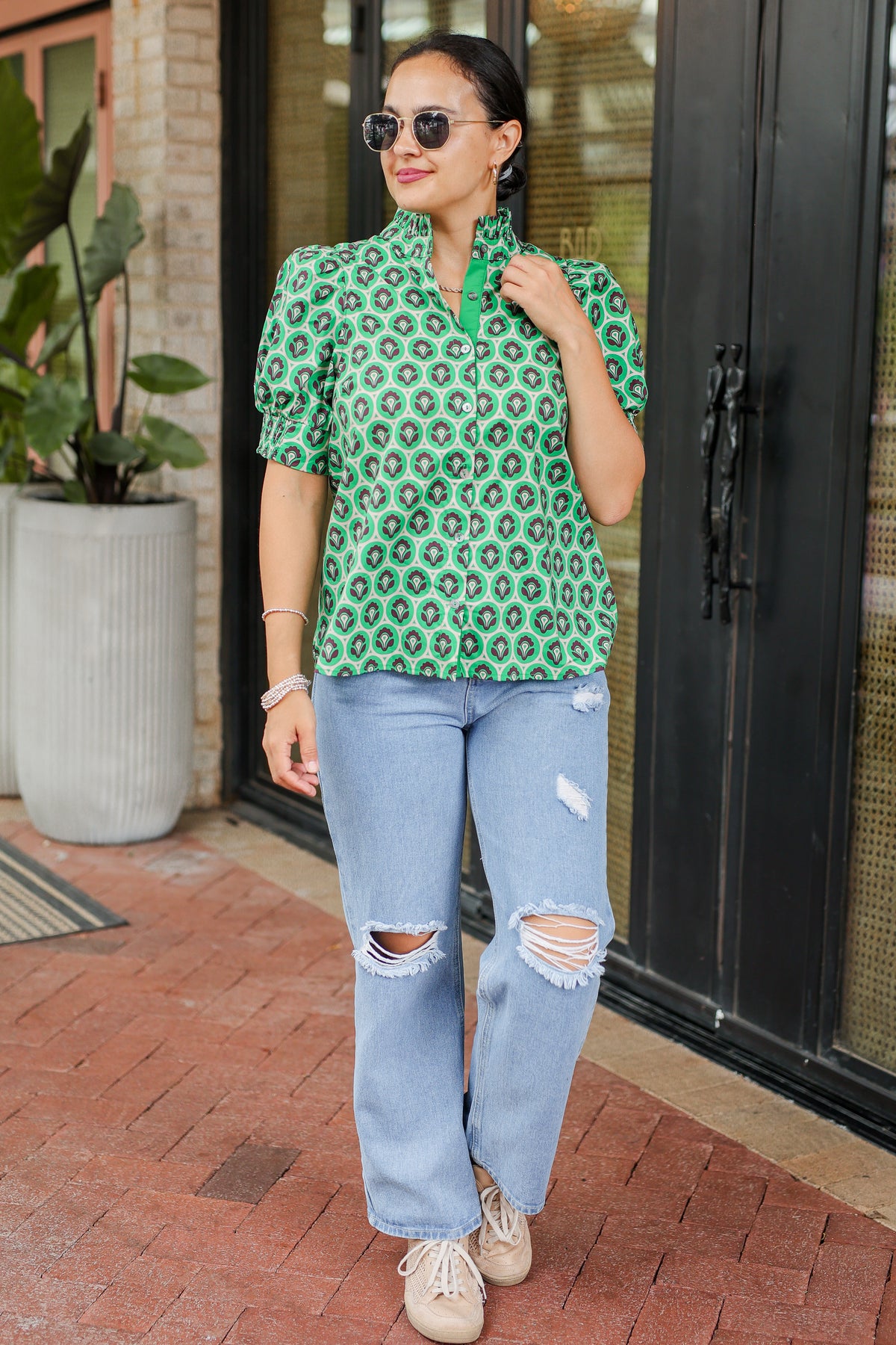 Woman wearing a green patterned blouse and blue jeans standing on a brick patio.