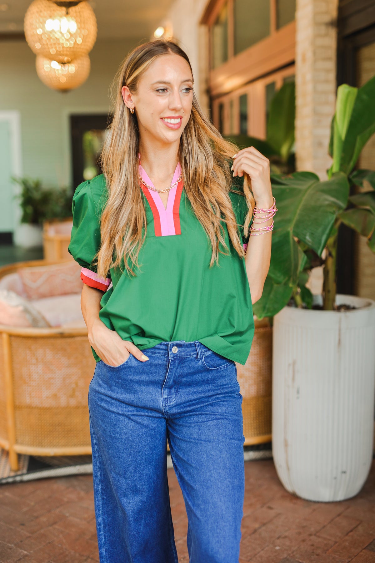 Woman in a green top and blue jeans standing outdoors with plants and a building in the background