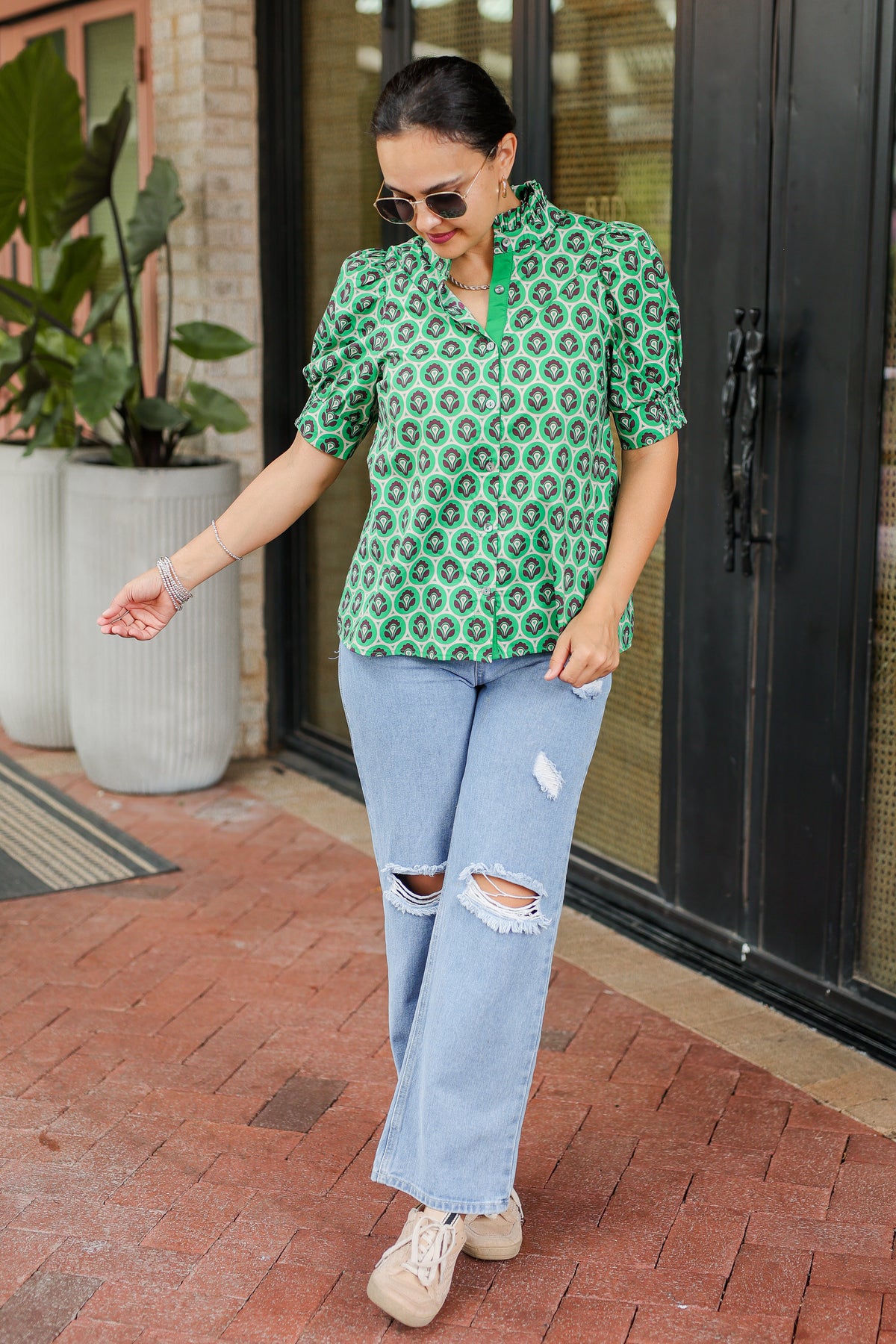 Woman wearing a green patterned shirt and blue jeans standing on a brick patio.