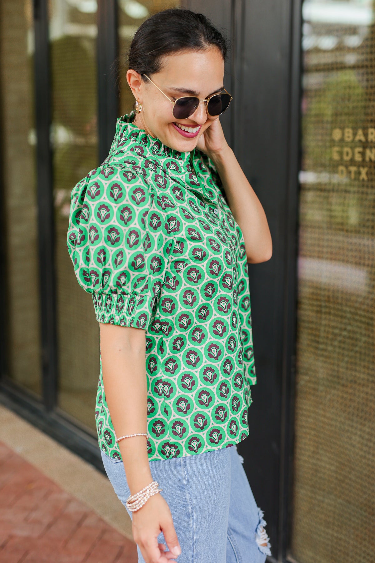 Woman wearing a green patterned blouse and sunglasses, standing in front of a store window.