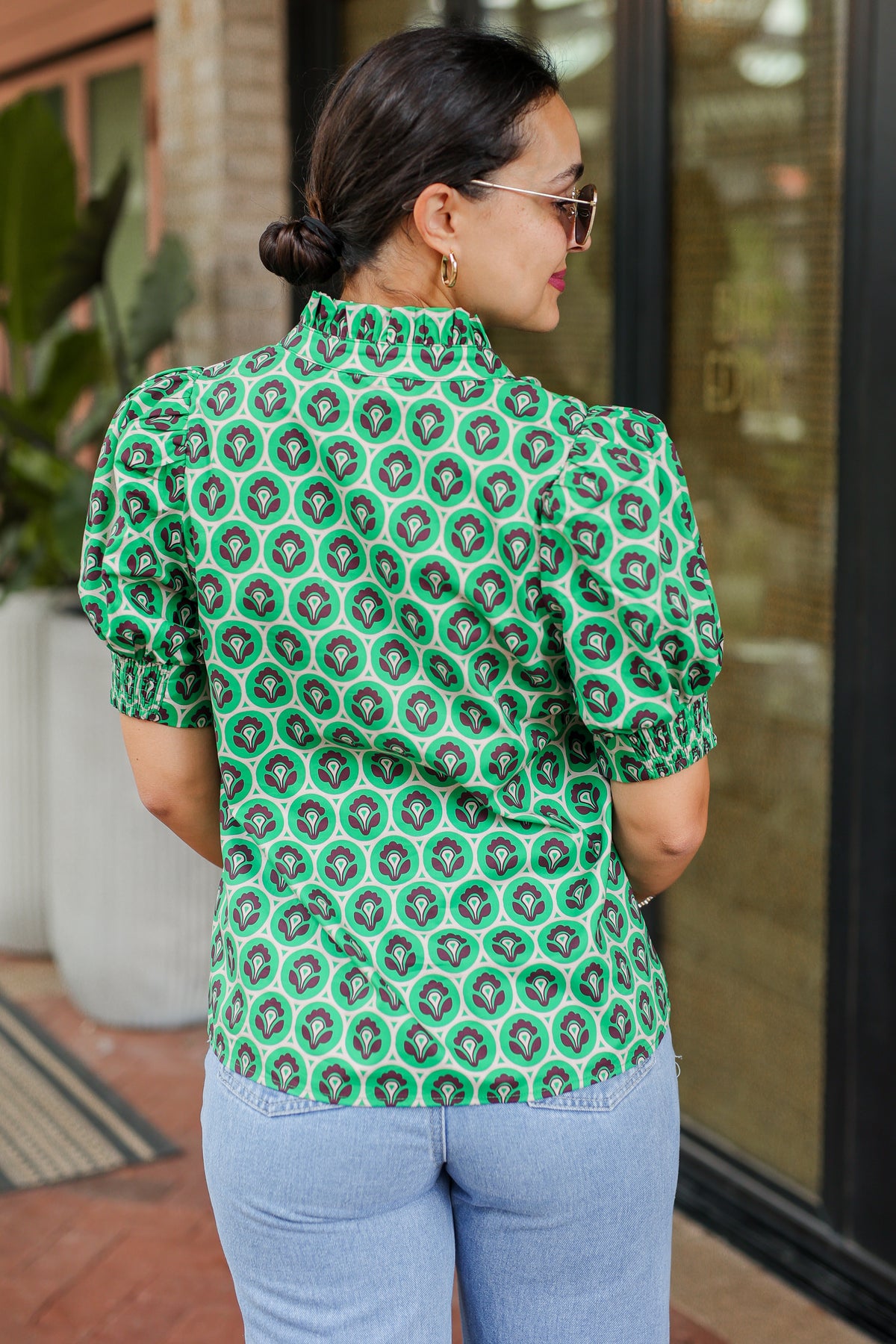 Woman wearing a green patterned blouse and blue jeans standing outdoors.