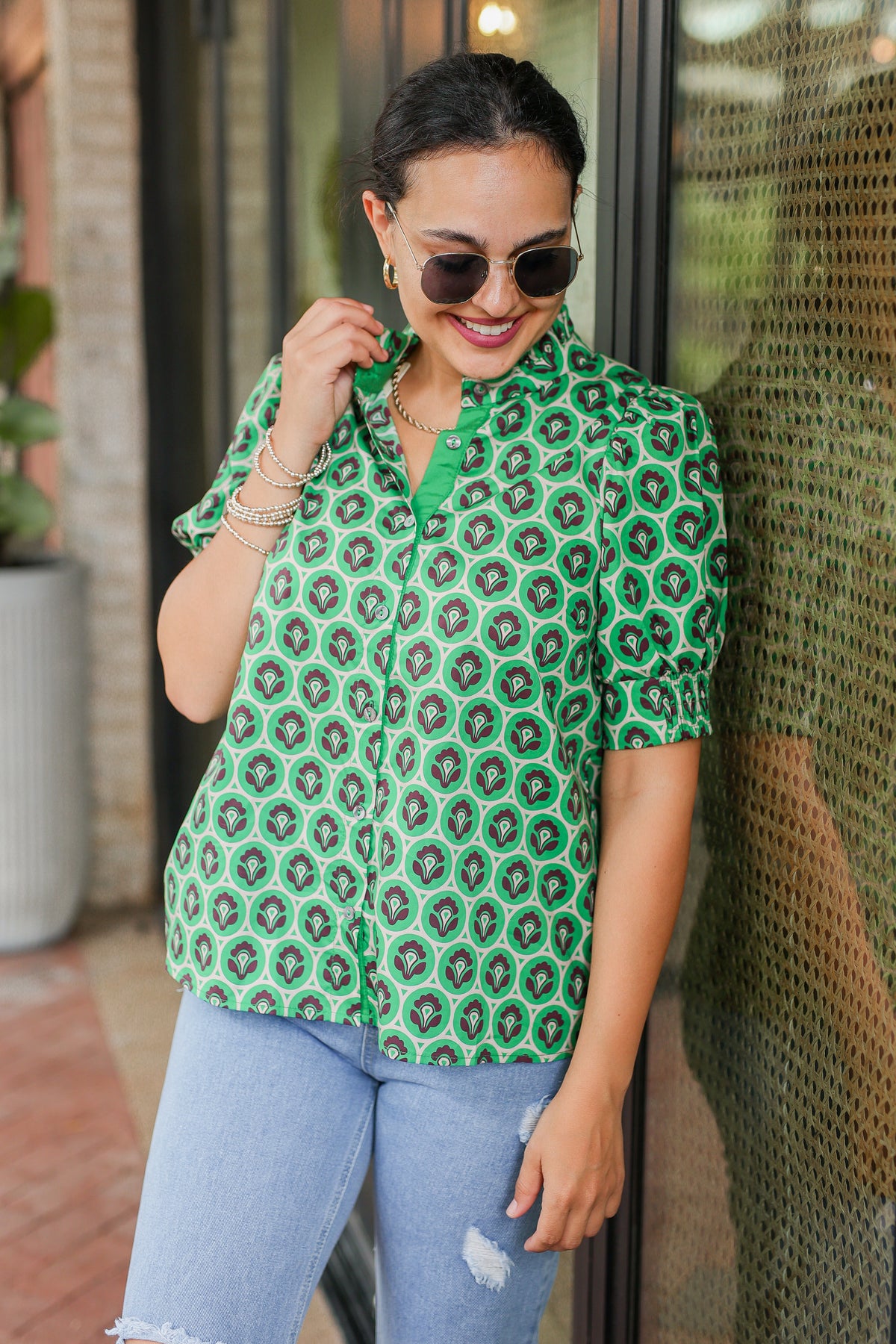 Woman wearing a green patterned shirt and sunglasses, standing against a textured wall.